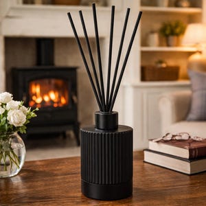 May include: A black reed diffuser with a ribbed design, containing black reeds. The diffuser is on a wooden table. A vase of white flowers and a fireplace are in the background. The room is warmly lit, creating a cozy atmosphere.