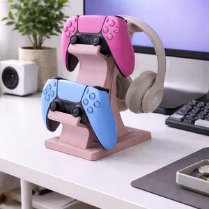 May include: A pink and light blue gaming controller stand holding a pink and a light blue controller, and a pair of gray headphones. The stand is on a white desk with a computer monitor in the background.