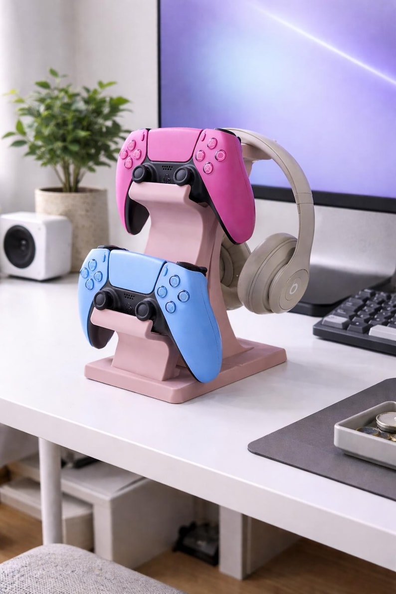 May include: A pink and light blue gaming controller stand holding a pink and a light blue controller, and a pair of gray headphones. The stand is on a white desk with a computer monitor in the background.