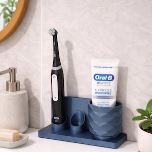 May include: A dark blue toothbrush holder with a geometric design, holding a black electric toothbrush and a tube of Oral-B 3D White toothpaste. The holder also has a small, round compartment. A modern bathroom accessory.