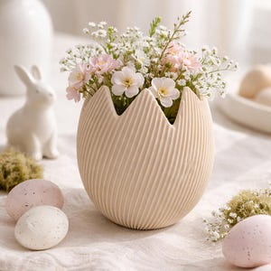 May include: A beige, egg-shaped vase with a ridged surface, filled with pink and white flowers. Decorative Easter eggs and a ceramic bunny are also visible, creating a spring theme.