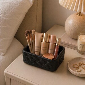 K&ouml;nnte beinhalten: Ein schwarzer, strukturierter Make-up-Organizer gef&uuml;llt mit Make-up-Pinseln, Foundation-Flaschen, Concealer und Lipgloss. Der Organizer steht auf einem Nachttisch neben einer Lampe, einer Kerze und einer kleinen Schale mit Schmuck.