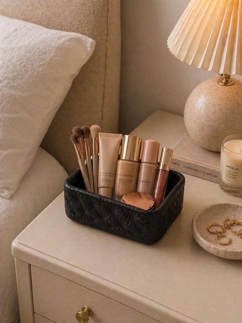 K&ouml;nnte beinhalten: Ein schwarzer, strukturierter Make-up-Organizer gef&uuml;llt mit Make-up-Pinseln, Foundation-Flaschen, Concealer und Lipgloss. Der Organizer steht auf einem Nachttisch neben einer Lampe, einer Kerze und einer kleinen Schale mit Schmuck.