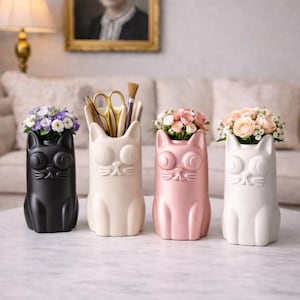 Könnte beinhalten: Vier katzenförmige Vasen in Schwarz, Creme, Rosa und Weiß, jede mit Blumen oder Schreibwaren. Die Vasen haben große, runde Augen und ein schlichtes, stilisiertes Design. Sie stehen auf einem Tisch mit Marmorplatte.
