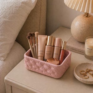 K&ouml;nnte beinhalten: Ein rosa, strukturiertes Make-up-Organizer gef&uuml;llt mit Make-up-Pinseln, Foundation, Concealer und Lipgloss. Der Organizer steht auf einem Nachttisch neben einer Lampe und einer kleinen Schale mit Schmuck. Eine brennende Kerze ist ebenfalls sichtbar.