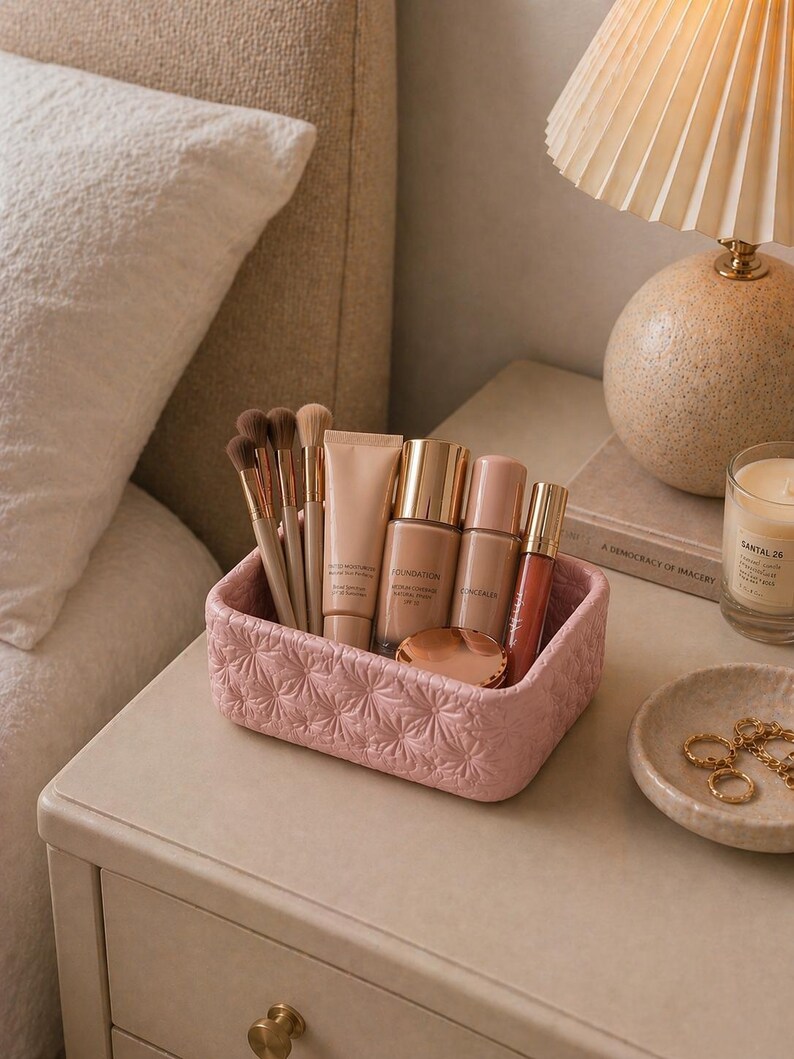 K&ouml;nnte beinhalten: Ein rosa, strukturiertes Make-up-Organizer gef&uuml;llt mit Make-up-Pinseln, Foundation, Concealer und Lipgloss. Der Organizer steht auf einem Nachttisch neben einer Lampe und einer kleinen Schale mit Schmuck. Eine brennende Kerze ist ebenfalls sichtbar.