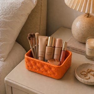 K&ouml;nnte beinhalten: Ein orangefarbener, strukturierter Make-up-Organizer gef&uuml;llt mit Make-up-Pinseln, Foundation, Concealer und Lipgloss. Der Organizer steht auf einem Nachttisch neben einer Lampe, einer Kerze und einer kleinen Schale mit Schmuck.