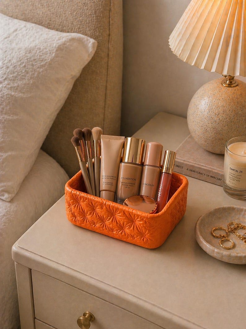 K&ouml;nnte beinhalten: Ein orangefarbener, strukturierter Make-up-Organizer gef&uuml;llt mit Make-up-Pinseln, Foundation, Concealer und Lipgloss. Der Organizer steht auf einem Nachttisch neben einer Lampe, einer Kerze und einer kleinen Schale mit Schmuck.