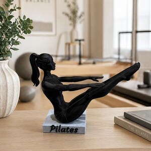 Peut inclure: Sculpture noire repr&eacute;sentant une personne en position de Pilates. La figurine est pos&eacute;e sur un socle rectangulaire avec le mot "Pilates" en blanc. La sculpture est sur une surface en bois clair.