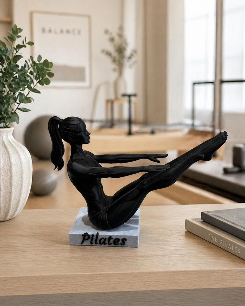 Peut inclure: Sculpture noire repr&eacute;sentant une personne en position de Pilates. La figurine est pos&eacute;e sur un socle rectangulaire avec le mot "Pilates" en blanc. La sculpture est sur une surface en bois clair.