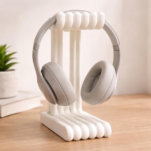 May include: A white plastic headphone stand with a curved base and vertical supports, holding a pair of grey over-ear headphones. The stand is designed to cradle the headphones, keeping them organised and accessible. The headphones have large, cushioned earcups.