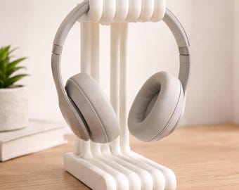 Peut inclure: Support de casque en plastique blanc avec une base incurvée et des supports verticaux, tenant une paire de casques circum-auriculaires gris. Le support est conçu pour maintenir les écouteurs, les gardant organisés et accessibles. Les écouteurs ont de grands coussinets.