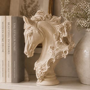 May include: A cream-coloured horse head bust sculpture featuring intricate, swirling mane details and a pedestal base. The sculpture is positioned next to three books titled Modern Home Decor, Beautiful Interiors, and The Styled Home.
