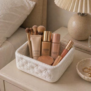 K&ouml;nnte beinhalten: Ein wei&szlig;er, gesteppter Korb mit Make-up-Produkten, darunter Foundation-Fl&auml;schchen, Pinsel und Lipgloss. Die Produkte sind auf einem cremefarbenen Nachttisch neben einer Lampe und einer kleinen dekorativen Schale angeordnet.