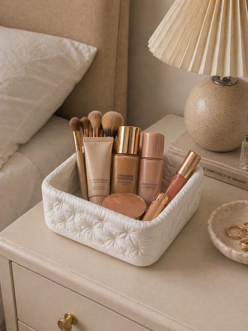 K&ouml;nnte beinhalten: Ein wei&szlig;er, gesteppter Korb mit Make-up-Produkten, darunter Foundation-Fl&auml;schchen, Pinsel und Lipgloss. Die Produkte sind auf einem cremefarbenen Nachttisch neben einer Lampe und einer kleinen dekorativen Schale angeordnet.