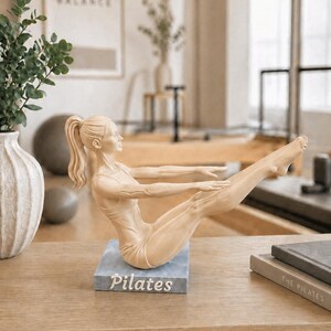 Peut inclure: Une figurine de Pilates beige dans une pose assise, les bras tendus et les jambes lev&eacute;es. La figurine est sur une base bleu-gris avec le mot "Pilates" en blanc. La figurine est sur une surface en bois.