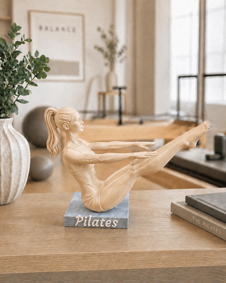 Peut inclure: Une figurine de Pilates beige dans une pose assise, les bras tendus et les jambes lev&eacute;es. La figurine est sur une base bleu-gris avec le mot "Pilates" en blanc. La figurine est sur une surface en bois.