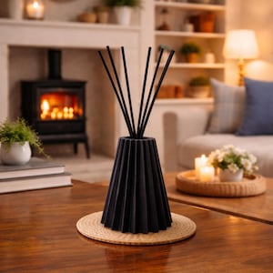 Könnte beinhalten: Ein schwarzer Reed-Diffusor mit schwarzen Stäbchen, auf einem gewebten Untersetzer, auf einem Holztisch. Der Diffusor hat ein plissiertes Design. Der Hintergrund zeigt einen Kamin, Pflanzen und ein Sofa.