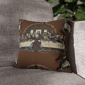 May include: A brown square cushion with a vintage depiction of The Last Supper. The design includes the text "THIS IS MY BODY, BROKEN FOR YOU" and "LUKE 22:19". A red heart is also featured in the artwork.