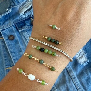 Op de afbeelding: Een collectie van vijf armbanden om de pols. Eén is een dunne goudkleurige ketting met heldere kristallen. De andere vier zijn ontwerpen van wikkeldraad met groene stenen kralen, waarvan er één ook een witte parelachtige kraal bevat.