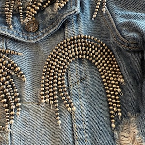 May include: Close-up of a light blue denim jacket adorned with silver beaded embellishments. The jacket features a distressed design with frayed edges and a button closure. The text "These are not finished bracelets" is visible.