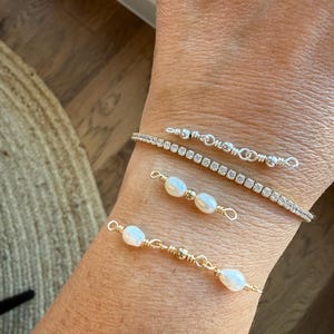 Peut inclure: Un ensemble de trois bracelets portés au poignet. L'un est une chaîne argentée avec de petites perles, un autre est un bracelet tennis avec des pierres claires, et deux sont des bracelets en fil doré avec des perles ovales blanches.