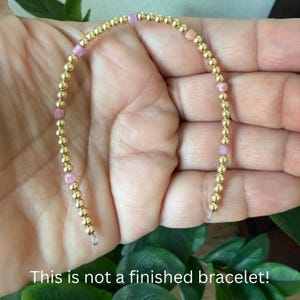 May include: A string of small, round, metallic gold-colored beads interspersed with occasional translucent pink beads. The beads are arranged in a curved line, and the text at the bottom of the image states: This is not a finished bracelet.
