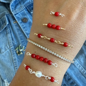 Peut inclure: Une collection de six bracelets portés sur le bras. Cinq bracelets présentent des perles rouges ou une perle blanche unique accentuée par un fil d'or ou d'argent. Un bracelet est une fine bande flexible sertie de cristaux clairs.