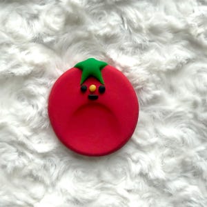 May include: A red tomato-shaped decoration with a green stem and a smiling face. The tomato has a yellow dot for a nose and black dots for eyes. The decoration is on a white, fluffy background.