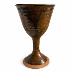 May include: A handmade, brown ceramic goblet with a wide, conical bowl and a slender stem. The goblet has a textured surface and a dark brown glaze on the upper half, with a lighter brown base. The base has a circular, unglazed rim.