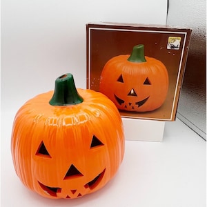 May include: An orange ceramic jack-o'-lantern with a green stem and carved triangular eyes and a smiling mouth. A second jack-o'-lantern is visible in a brown box. The Halloween decoration is a festive item for the fall season.
