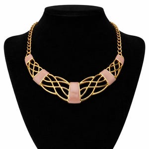 May include: A gold-toned necklace with a woven design and blush pink accents. The necklace is displayed on a black mannequin, showcasing its intricate details and elegant style. The chain is a classic curb link.