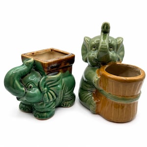 May include: Two ceramic elephant-shaped planters with glossy finishes. The left planter is green with a square brown rim. The right planter features a green elephant holding a cylindrical tan container with vertical ridges and a smooth brown rim.
