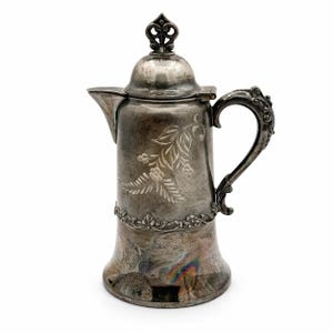 May include: Antique silver-plated coffee pot with a decorative floral design. The pot features an ornate handle and a detailed lid finial. The body of the pot is engraved with a floral pattern and a decorative band near the base.