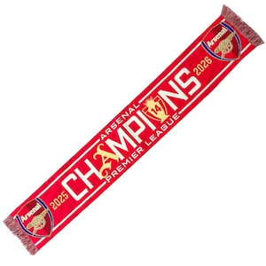 May include: A red knit scarf featuring the Arsenal football club crest at both ends. The text reads Arsenal Champions Premier League with the years 2025 and 2026, and a gold trophy icon with the number 14. The scarf has fringed ends in red and white.