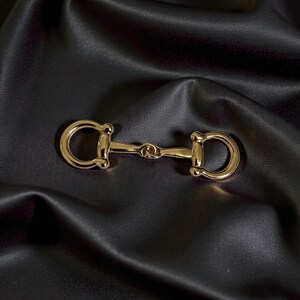 May include: A polished gold-tone metal horsebit hardware piece featuring a central bar connected to two rounded D-shaped rings on either side. The hardware has a smooth, reflective surface and a classic equestrian-inspired design shape.