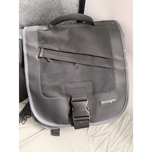 Peut inclure: Sacoche noire avec une poignée supérieure, une bandoulière et un rabat avant fixé par une boucle. Le sac a un corps en tissu texturé avec une garniture en similicuir et la marque "Kensington" est visible.