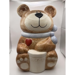 Op de afbeelding: Een keramische koekjespot in de vorm van een bruine teddybeer. De beer heeft een wit gezicht, een zwarte neus en een lichtblauwe sjaal. Een rood hart zit op de borst van de beer en op de pot staat "COOKIES".