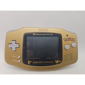 May include: A gold-colored Game Boy Advance handheld console. The console features a black screen, white buttons, and the text "Pokémon" and "Game Boy Advance". The console also has the text "Pokémon Center NEW YORK".