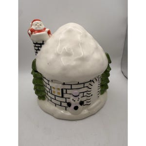 May include: A ceramic Christmas house figurine with a snowy roof and brick detailing. A Santa Claus figure is perched on the chimney. Green trees flank the sides, and a snowman face is on the front. The house has a small window.