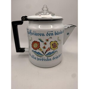 Puede incluir: Cafetera de esmalte blanco con asa y boquilla negras. La cafetera presenta un diseño floral en rojo, amarillo y azul, con texto en escritura azul. La tapa es de cristal transparente con un pomo decorativo.