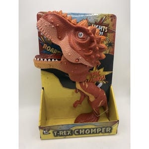 May include: A red and orange T-Rex Chomper toy with a detailed, textured design. The toy is in a yellow box with the text "LIGHTS!" and "EPIC SOUNDS!" on the packaging. The dinosaur's mouth is open, revealing white teeth.