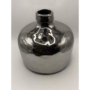 May include: A silver-colored vase with a rounded body and a narrow neck. The vase has a shiny, metallic finish that reflects light. The opening at the top is dark. This decorative vase is suitable for home decor.