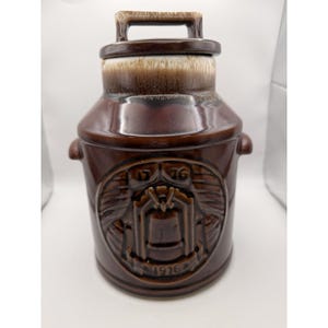 May include: A brown ceramic cookie jar with a lid and handle. The jar features a raised emblem with the numbers "1776" and "1976". The jar has a glossy finish and a cylindrical shape, with a slightly lighter brown color on the lid.