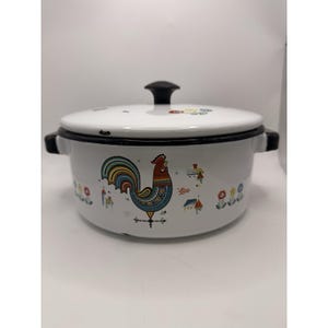 Puede incluir: Olla de esmalte blanco con asa y tapa negras. La olla presenta un colorido diseño de gallo con cola azul, amarilla y roja. También hay ilustraciones adicionales de flores y casas.