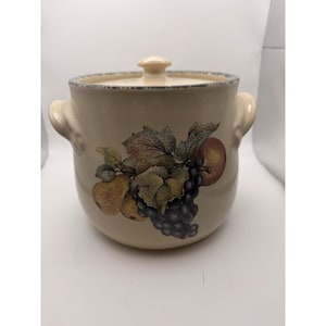 Puede incluir: Olla de cerámica beige con tapa y dos asas laterales. La olla presenta un diseño de frutas con manzanas, uvas y peras. La tapa y el borde son de color beige claro. La olla es redonda y tiene una superficie texturizada.
