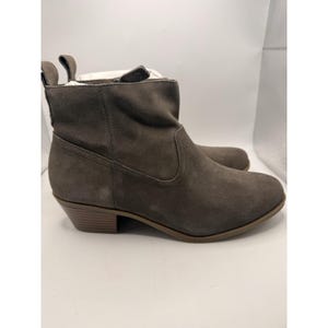 以下が含まれることがあります： 低いヒールが付いたグレーのスエードアンクルブーツのペア。ブーツは丸いつま先と背面にプルタブが付いています。ヒールの高さは約5cmです。