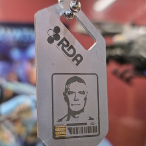 Puede incluir: Un pendrive USB de metal plateado con forma de chapa de identificación, con una cadena de bolas. La chapa presenta el logotipo de RDA, un retrato y un código de barras. El conector USB es visible en la parte inferior.