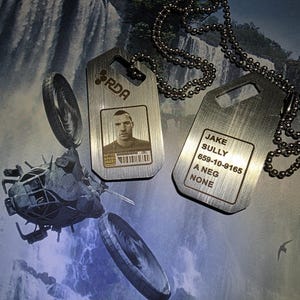 Puede incluir: Dos placas de identificación plateadas con collares de cadena de bolas. Una placa presenta una foto y las letras "RDA". La otra placa tiene el nombre "Jake Sully", un número de teléfono y otro texto. Un vehículo futurista y una cascada de fondo son visibles.