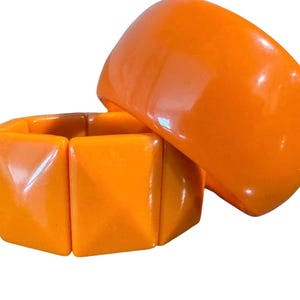 May include: Two orange bracelets. One is a wide, curved bangle. The other is a stretch bracelet made of square, glossy, orange beads. The bracelets are set against a white background.